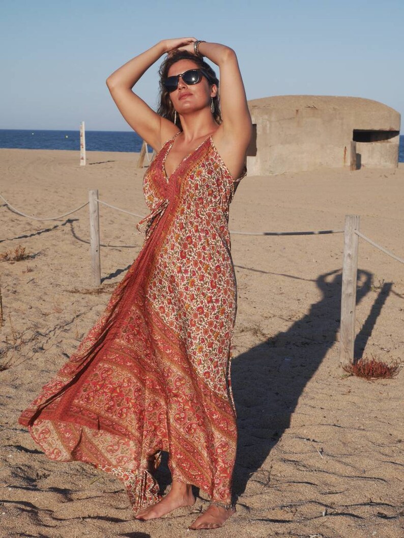May include: A woman wearing a long, red and white floral print dress with a halter neckline. She is standing on a sandy beach with a concrete bunker in the background.