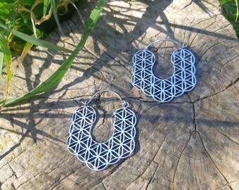 May include: A pair of silver hoop earrings featuring a geometric flower of life design. The earrings are displayed on a wooden surface.