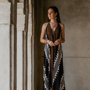 May include: A woman wearing a black and white patterned halter dress with orange accents stands in a doorway. She is barefoot and has long dark hair. The doorway is made of stone and has a textured surface.