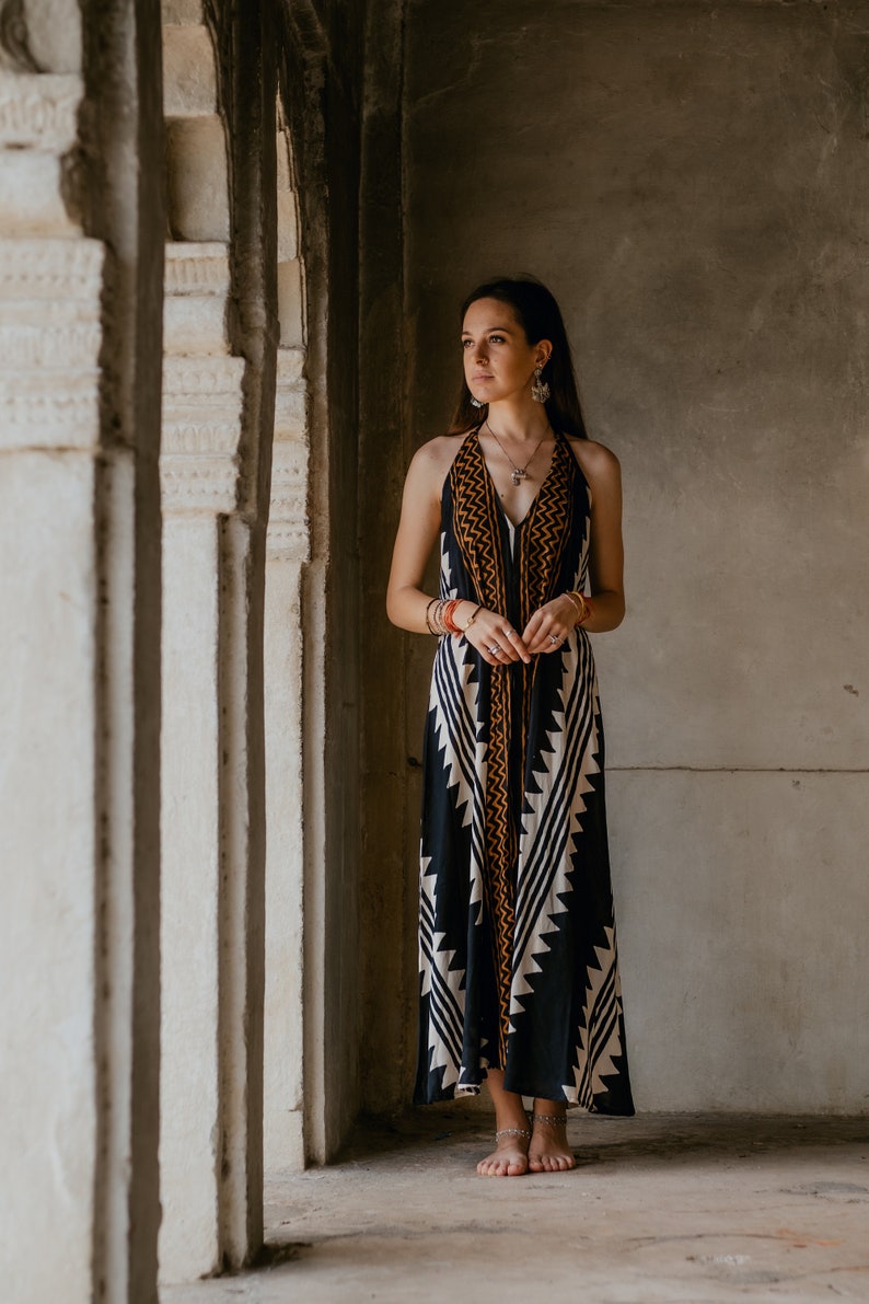 May include: A woman wearing a black and white patterned halter dress with orange accents stands in a doorway. She is barefoot and has long dark hair. The doorway is made of stone and has a textured surface.