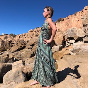 Könnte beinhalten: Eine Frau in einem langen, fließenden, grün-schwarz gemusterten Kleid steht an einem felsigen Strand. Das Kleid hat ein Paisley-Muster und einen V-Ausschnitt. Die Frau schaut nach rechts und ihre Haare sind vom Gesicht zurückgezogen.