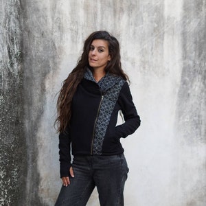 K&ouml;nnte beinhalten: Eine schwarze Rei&szlig;verschlussjacke mit einem geometrischen Muster auf der rechten Seite. Die Jacke hat einen hohen Kragen und lange &Auml;rmel.