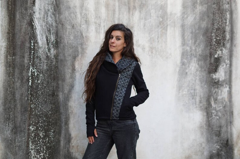 K&ouml;nnte beinhalten: Eine schwarze Rei&szlig;verschlussjacke mit einem geometrischen Muster auf der rechten Seite. Die Jacke hat einen hohen Kragen und lange &Auml;rmel.