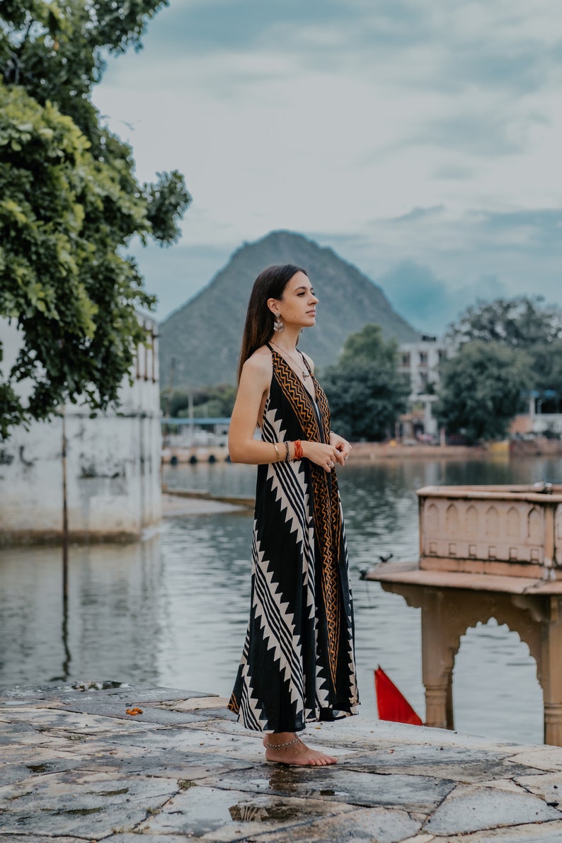 May include: A woman wearing a black and white patterned maxi dress with a brown and gold trim stands on a stone platform by a body of water. She is looking off to the side, and there is a hill in the background.