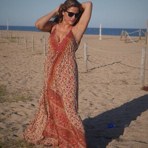 May include: A woman wearing a long, flowing maxi dress with a floral print in shades of red, orange, and white. The dress has a deep V-neckline and a relaxed fit. She is standing on a sandy beach with a blue sky and ocean in the background.