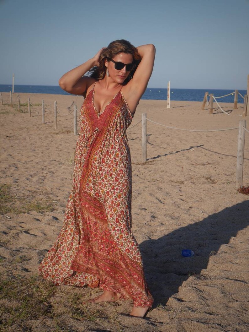 May include: A woman wearing a long, flowing maxi dress with a floral print in shades of red, orange, and white. The dress has a deep V-neckline and a relaxed fit. She is standing on a sandy beach with a blue sky and ocean in the background.