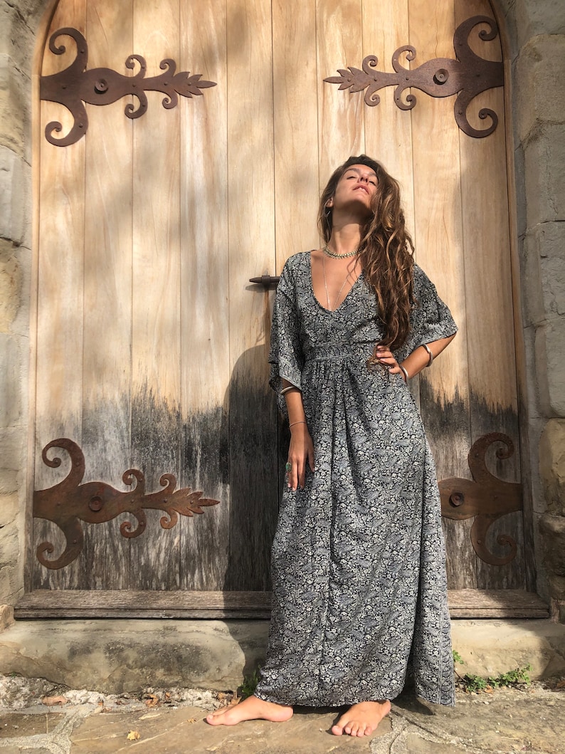 May include: A woman wearing a long, black and white floral print dress with a V-neck and 3/4 sleeves. She is standing in front of a wooden door with iron hinges.