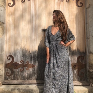 May include: A woman wearing a long, black and white floral print dress with a deep V-neckline. She is standing in front of a wooden door with ornate iron hinges.