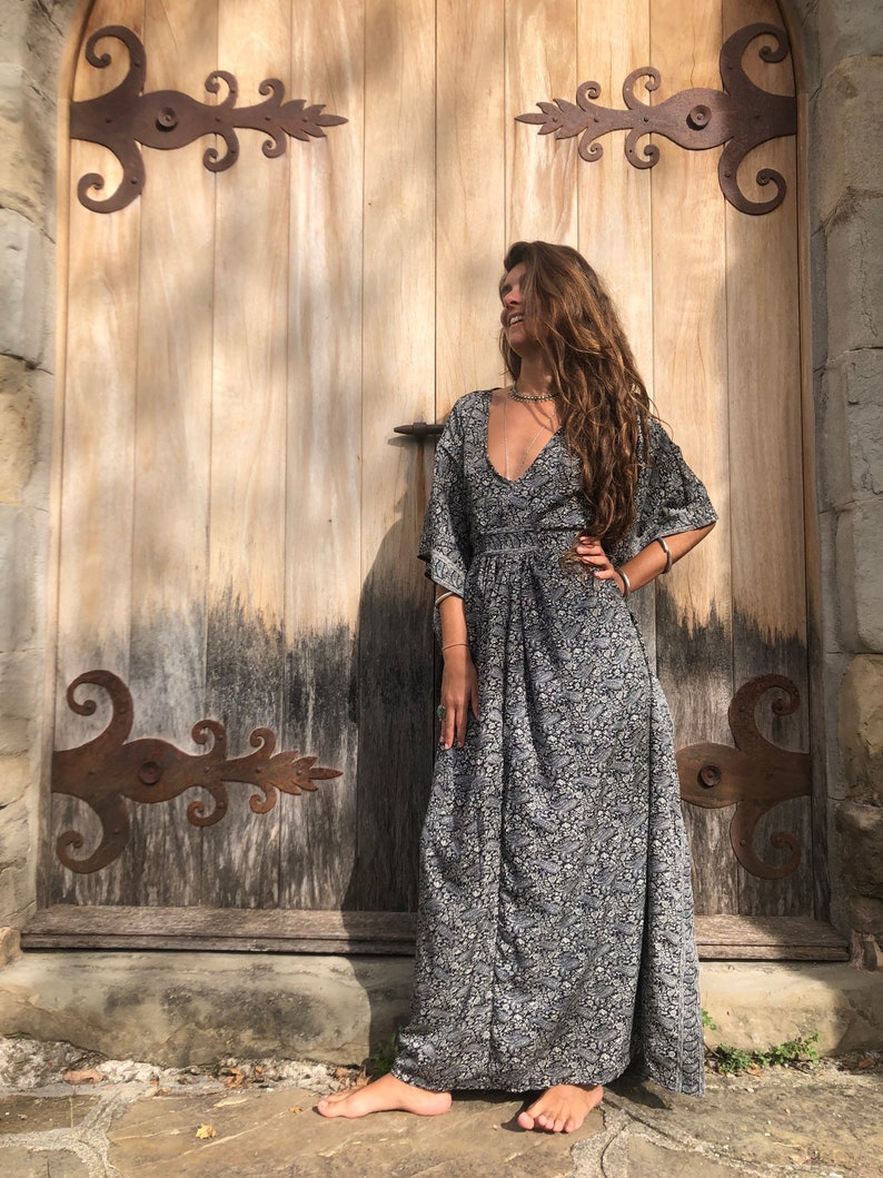 May include: A woman wearing a long, black and white floral print dress with a deep V-neckline. She is standing in front of a wooden door with ornate iron hinges.