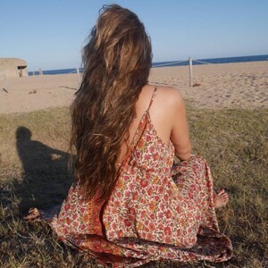 May include: A woman with long brown hair sits on the grass wearing a floral maxi dress. The dress has a red, orange, and yellow floral pattern on a white background. The woman is facing away from the camera and looking out at the sea.