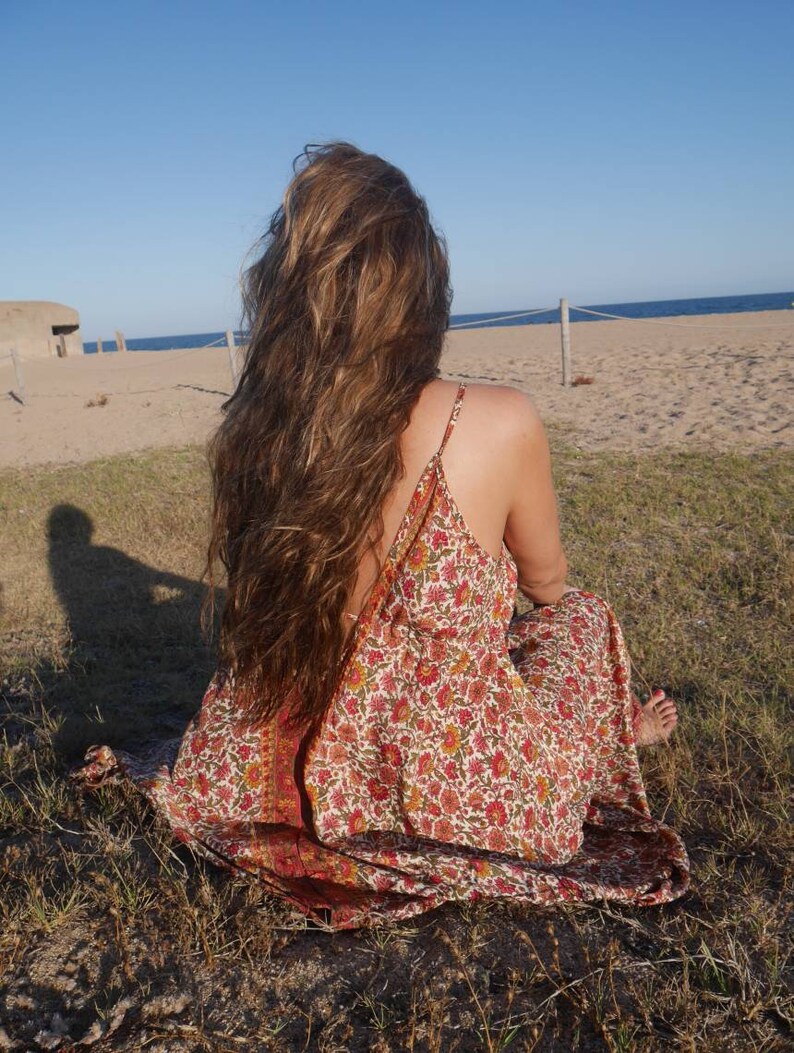 May include: A woman with long brown hair sits on the grass wearing a floral maxi dress. The dress has a red, orange, and yellow floral pattern on a white background. The woman is facing away from the camera and looking out at the sea.