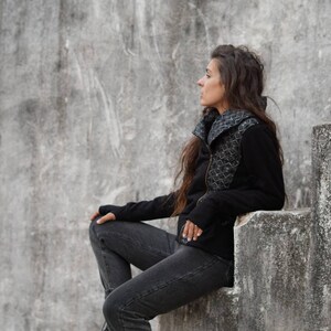 K&ouml;nnte beinhalten: Eine Frau in einer schwarzen Jacke mit geometrischem Muster sitzt auf einer Betontreppe. Sie schaut zur Seite und hat lange braune Haare.
