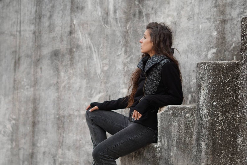 K&ouml;nnte beinhalten: Eine Frau in einer schwarzen Jacke mit geometrischem Muster sitzt auf einer Betontreppe. Sie schaut zur Seite und hat lange braune Haare.