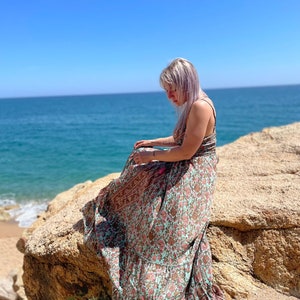May include: A woman wearing a long, flowing floral maxi dress with a light blue and pink color scheme. She is sitting on a large rock overlooking a blue ocean with a sandy beach in the background.