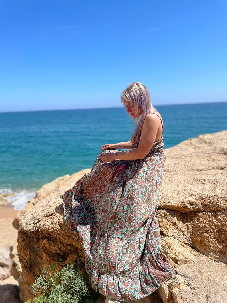 May include: A woman wearing a long, flowing floral maxi dress with a light blue and pink color scheme. She is sitting on a large rock overlooking a blue ocean with a sandy beach in the background.