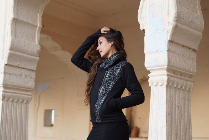 K&ouml;nnte beinhalten: Eine schwarze Rei&szlig;verschlussjacke mit einem silbernen und schwarzen Muster auf der linken Seite. Die Jacke hat einen hohen Kragen und einen Rei&szlig;verschluss.