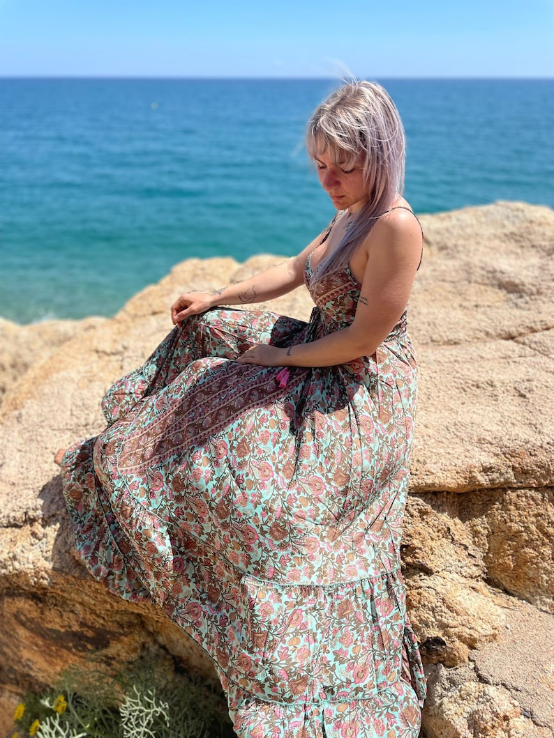 May include: A woman wearing a long, flowing floral dress with a light blue background and brown and pink floral patterns. She is sitting on a rocky cliff overlooking the ocean.