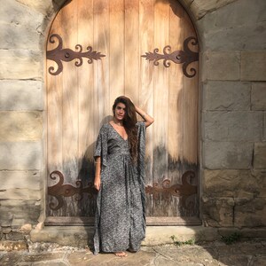 May include: A woman wearing a long, gray and white patterned dress stands in front of a wooden door with ornate iron hinges. The door is set in a stone archway.