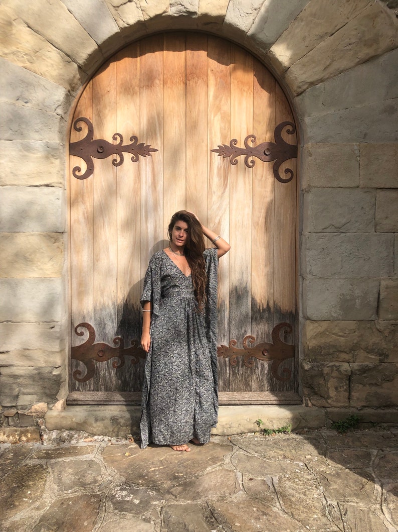 May include: A woman wearing a long, gray and white patterned dress stands in front of a wooden door with ornate iron hinges. The door is set in a stone archway.