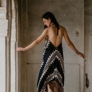 May include: A woman wearing a black and white patterned maxi dress with a halter neckline. The dress has a geometric design and a brown and orange trim at the bottom. She is standing in a doorway with a white wall behind her.