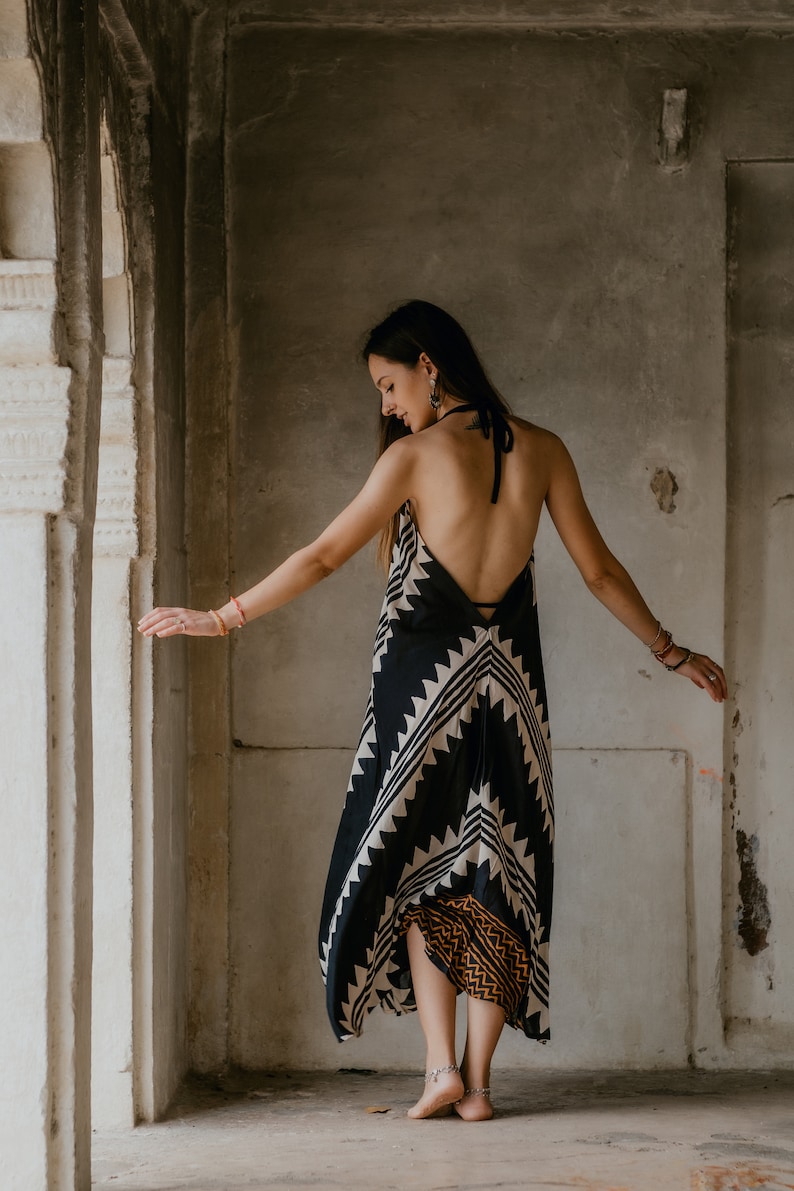 May include: A woman wearing a black and white patterned maxi dress with a halter neckline. The dress has a geometric design and a brown and orange trim at the bottom. She is standing in a doorway with a white wall behind her.