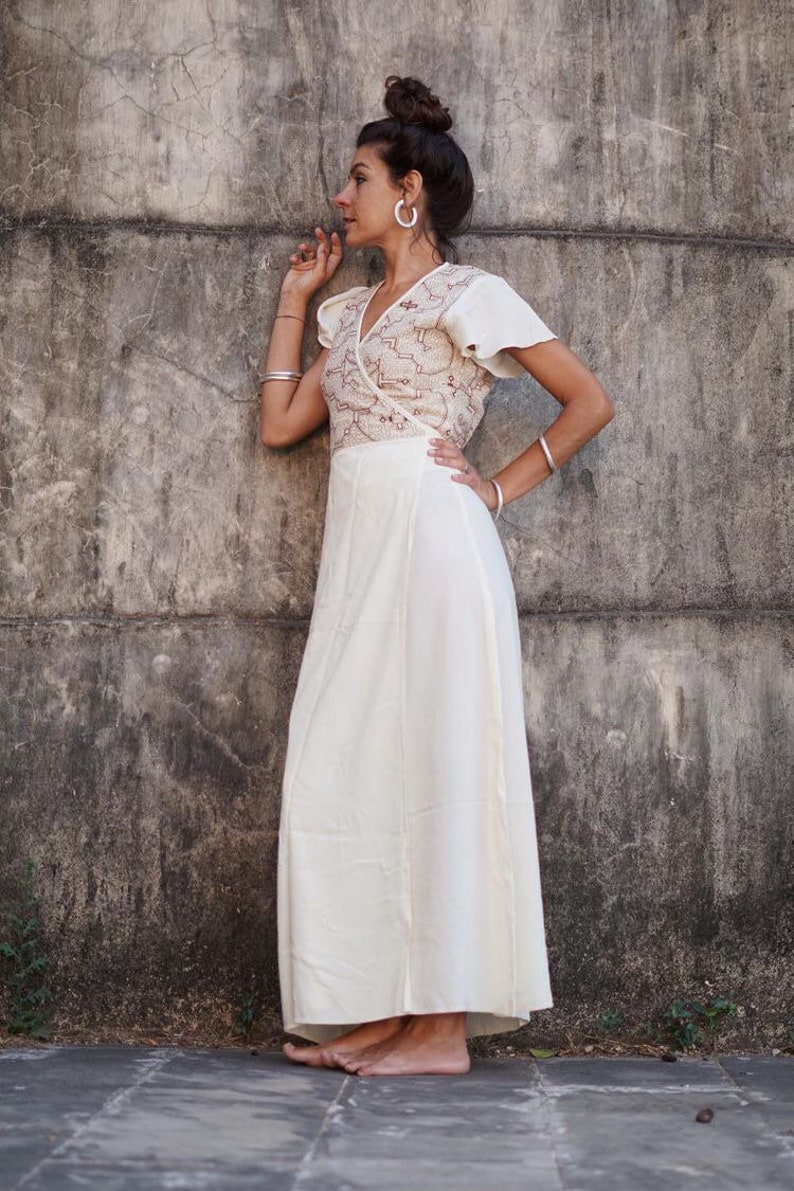 Peut inclure: Une robe blanche enroul&eacute;e avec un motif brod&eacute; brun. La robe a une manche courte et une jupe fluide. Le mannequin est debout devant un mur en b&eacute;ton.
