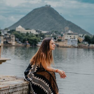 May include: A woman wearing a black and white patterned dress sits on stone steps overlooking a body of water. A large hill is in the background.