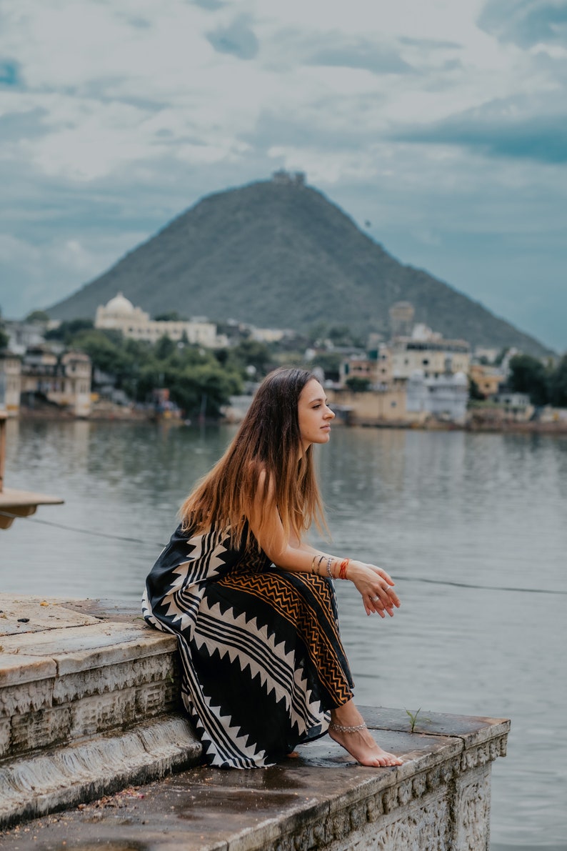 May include: A woman wearing a black and white patterned dress sits on stone steps overlooking a body of water. A large hill is in the background.