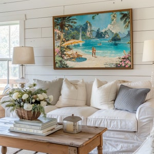 May include: A white couch with white and gray pillows sits in front of a white shiplap wall. A framed painting of a tropical beach scene hangs above the couch. A wooden coffee table with books and a candle lantern sits in front of the couch.