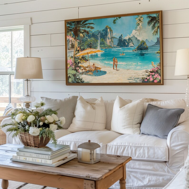 May include: A white couch with white and gray pillows sits in front of a white shiplap wall. A framed painting of a tropical beach scene hangs above the couch. A wooden coffee table with books and a candle lantern sits in front of the couch.