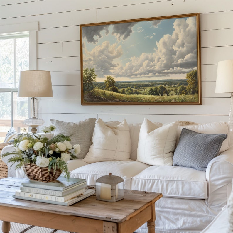 May include: A white sofa with white and gray pillows sits in front of a white shiplap wall with a framed landscape painting above it. A wooden coffee table with books and a candle lantern sits in front of the sofa.