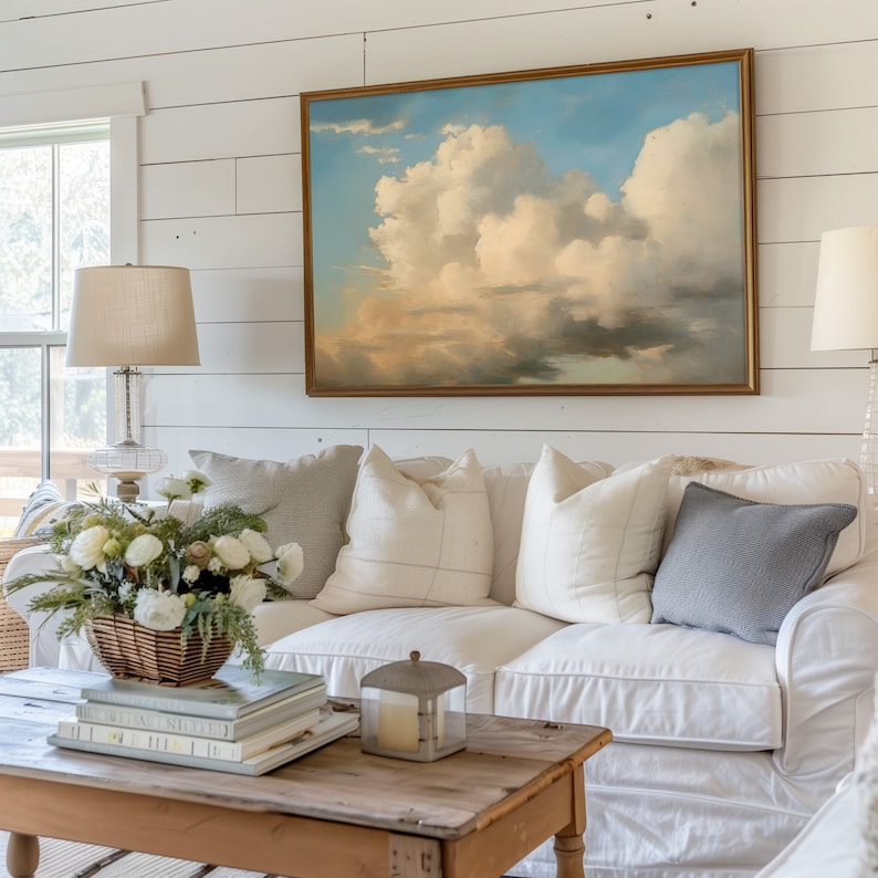 May include: A white couch with a variety of pillows, including a gray pillow, sits in front of a white shiplap wall. A framed painting of a cloudy sky hangs above the couch. A wooden coffee table sits in front of the couch with a stack of books and a candle lantern on top.