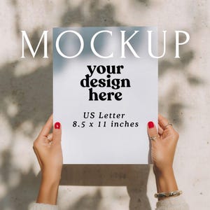May include: A white US Letter mockup with the text "MOCKUP" at the top and "your design here" in the center. The dimensions are listed as 8.5 x 11 inches. The paper is held by hands with red nail polish.