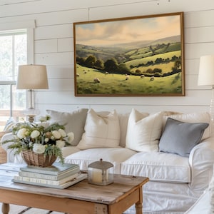 May include: A white sofa with a variety of pillows, including a gray pillow, sits in front of a white shiplap wall. A framed landscape painting of a green valley with sheep hangs above the sofa. A wooden coffee table with a stack of books and a lantern sits in front of the sofa.