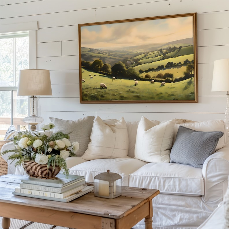 May include: A white sofa with a variety of pillows, including a gray pillow, sits in front of a white shiplap wall. A framed landscape painting of a green valley with sheep hangs above the sofa. A wooden coffee table with a stack of books and a lantern sits in front of the sofa.