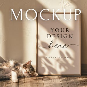 May include: A framed poster mockup with the word "MOCKUP" at the top. The poster has the text "YOUR DESIGN here" and is leaning against a wall. A sleeping tabby cat rests on the wooden floor in front of the poster.
