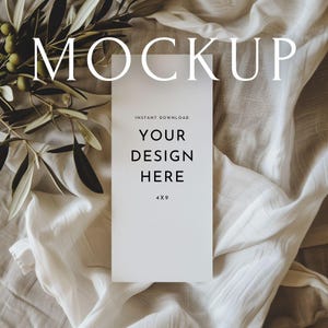 Puede incluir: Maqueta de tarjeta blanca de 4x9 pulgadas con el texto "MOCKUP" en la parte superior y "DESCARGA INSTANTÁNEA" y "YOUR DESIGN HERE" en el centro. Ramas de olivo con aceitunas verdes en la esquina superior izquierda. La tarjeta descansa sobre una tela blanca texturizada.