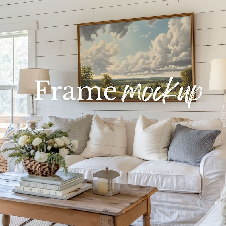 May include: A white couch with pillows and a throw blanket in a living room setting. A framed landscape painting hangs on the wall above the couch. A wooden coffee table sits in front of the couch with a stack of books and a candle lantern on top.