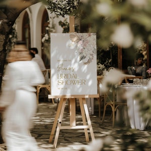 May include: A white sign with gold lettering on an easel, announcing "Welcome to Eleanor's Bridal Shower 18.06.23". The sign features a floral design with dried grasses and leaves.