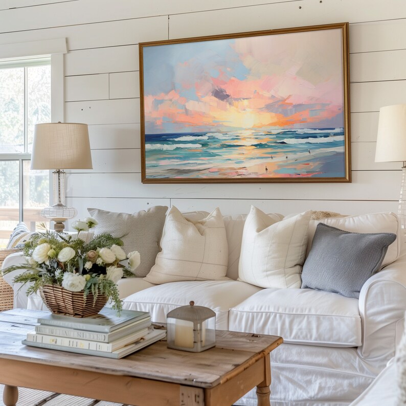 May include: A white couch with pillows in a living room setting. The couch is in front of a wall with white shiplap paneling. A framed painting of a seascape with a pink and orange sunset hangs on the wall above the couch.