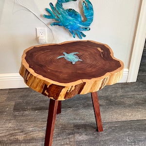 May include: A wooden side table with a live edge, featuring a turtle inlay. The table has three dark red legs. A blue and green crab wall art hangs above the table. The table is approximately 76 cm high.