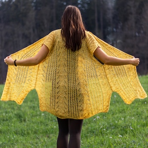 Puede incluir: Un cárdigan de punto amarillo con un patrón de encaje. El cárdigan está abierto por delante y tiene una forma larga y fluida.