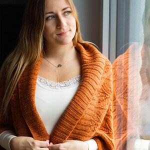 May include: A woman wearing a white vest top with a lace trim and a rust-coloured knitted cardigan. She is standing by a window and looking off to the side.