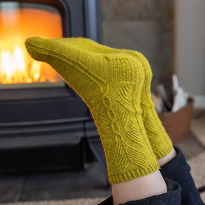 May include: A pair of yellow knitted socks with a cable knit pattern. The socks are being held up by a person's legs, with the feet resting on a rug in front of a fireplace.