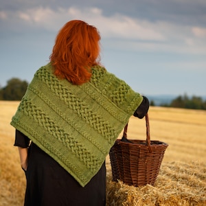 May include: A woman wearing a green knitted shawl with a braided pattern stands in a field of hay with a wicker basket.