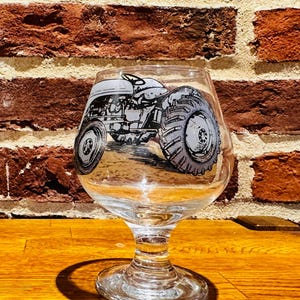 May include: A clear glass beer goblet with a detailed black and white illustration of a vintage tractor. The tractor design is printed on the front of the glass. The goblet has a stem and a wide, rounded bowl.