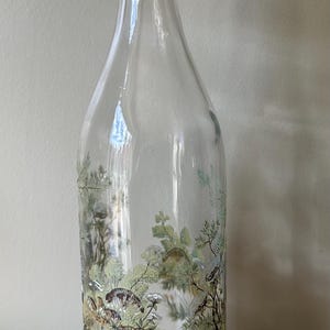 Puede incluir: Botella de vidrio transparente con cierre metálico y tapón rojo. La botella presenta una escena decorativa de bosque con árboles y animales en tonos verdes, marrones y beige. La botella está diseñada para líquidos.