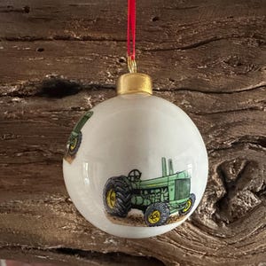 May include: White ceramic Christmas bauble featuring a green tractor design. The bauble has a gold cap and a red ribbon for hanging. The tractor has yellow wheels and is set against a white background.