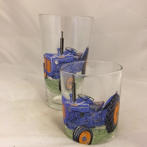 May include: Two glass tumblers with a blue tractor design. The tractor is blue with orange wheels and is set against a green field background.