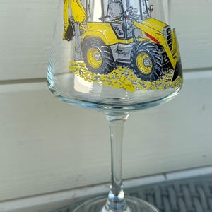 May include: A clear wine glass featuring a yellow and gray illustration of a backhoe loader. The construction vehicle is depicted with detailed tires and a yellow bucket. The glass has a long stem and a round base.